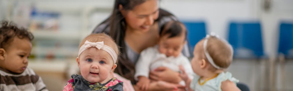 image of young babies being cared for in childcare setting. baby girl with blonde hair and pink headband with other babies the background